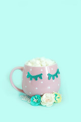 Pink mug with white polka dots with blue closed eyes with coffee and marshmallows and white, yellow and blue flowers nearby. Shiny eyelashes. Blue background..