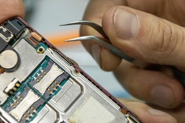 Elderly and young men repair a mobile phone together. They work in a repair shop. They use different tools. They are repair specialists.