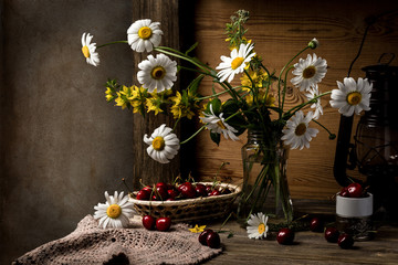 Still life with a bouquet of chamomile and lysimachia flowers in a glass jar, and cherries on the table.