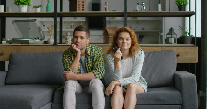 Young couple is watching TV, sitting on sofa at home, woman is interested, watching her favorite TV show, film, man is bored and uninterested. Caucasian, white. 4K, shot on RED camera.