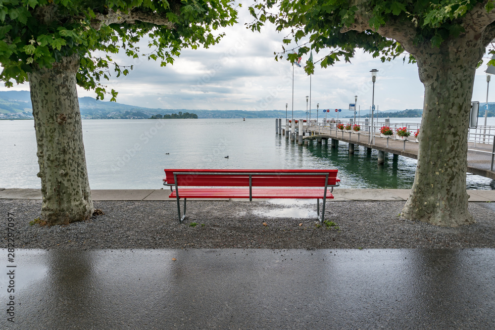 Wall mural red park bench on a lake shore with a view of the harbor pier and framed by london plane trees