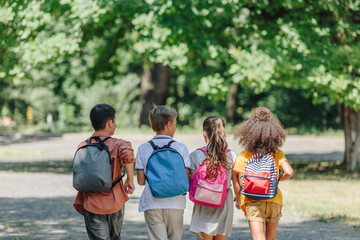 back view of four multicultural friends with backpacks running in summer park