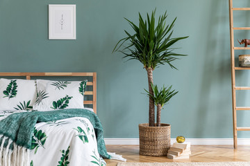Stylish interior design of bedroom with wooden bed, mock up poster frame, rattan ladder, tropical plants, books and elegant accessories. Beautiful bed sheets, blanket and pillow. Template. Home decor