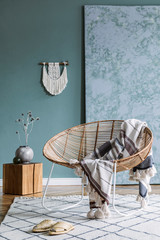Interior design of living room at modern home with stylish rattan armchair, plaid, macrame, wooden cube, flowers in vase, carpe and elegant accessories. Design home decor.Template. Abstract paintings