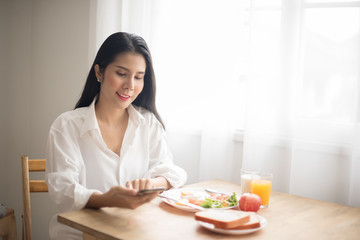 Obraz na płótnie Canvas Asian women in white shirt ordering products online with mobile phones in the morning. Happy woman with smartphone and breakfast on the table-Image