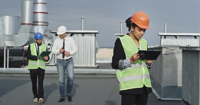 Old architect man and two engineers man and African woman analyzing the plan of construction site wearing the helmets on the rooftop of building commercial video