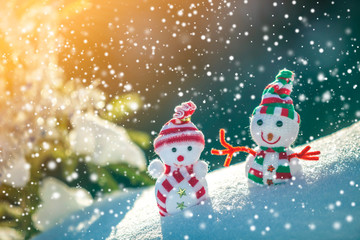 Two small funny toys baby snowman in knitted hats and scarves in deep snow outdoors near pine tree branch. Happy New Year and Merry Christmas greeting card.