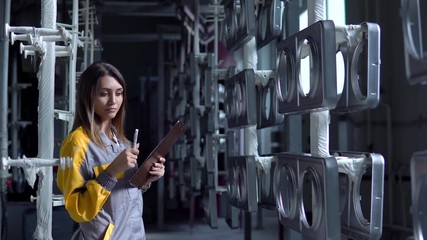 A young pretty white woman works in the paint shop of the plant, she checks the unpainted metall products and making record in check list.