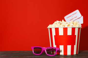 3d glasses, striped bucket with popcorn and tickets against red background