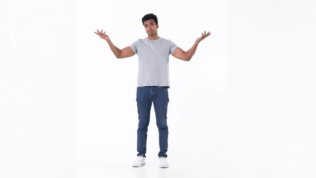 Portrait of puzzled and confused handsome young Indian man in plain blue jeans and grey t-shirt shrugging his shoulder on white background