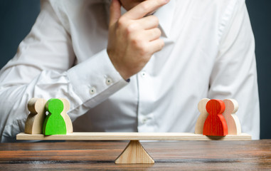 Man holds hand on chin looks at the rival red and green groups figures on a scales. Weighted...