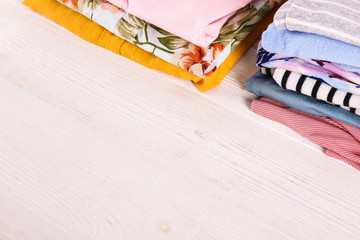 Stack of clean freshly laundered, neatly folded women's clothes on wooden table. Pile of shirts, dresses and sweaters on white board, concrete wall background. Copy space, close up, top view.