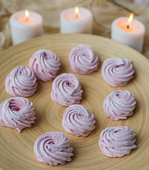 Merengue Marshmallow Zephyr lined in a circle on a bamboo plate on a white wooden background and three white burning candles. Top view. Pink sweet homemade zephyr or marshmallow.