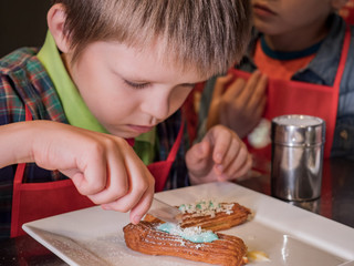 Funny children in red aprons eat eclairs cooked at master class. Confectionery education. Cookies, cakes, sweet cream, puff pastry. Vanilla, mint and confectionery powders