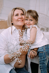 Gentle light portrait of mother and daughter. White clothes, a bright room, a sprig of gypsophila in his hands. Motherhood, love concept