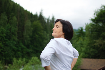 Senior woman doing a stretching exercise for the upper arms outside over landscape of forest and...