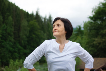 Senior woman doing a stretching exercise for the upper arms outside over landscape of forest and mountains