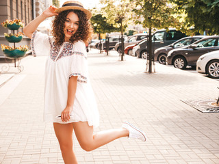 Portrait of beautiful smiling model with afro curls hairstyle dressed in summer hipster white dress.Sexy carefree girl posing in the street background.Trendy funny and positive woman in hat