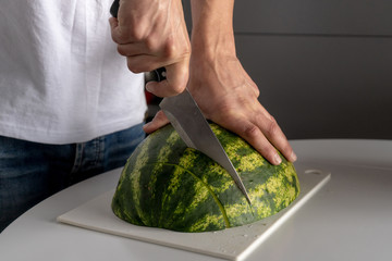 A man in a white T-shirt in the kitchen cuts with a knife half a juicy ripe sweet watermelon lying flesh down on a cutting board on the table.