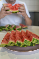 Sliced, juicy, ripe watermelon with black seeds on the plate is on the table. A man in a white T-shirt sits at a table in the kitchen and eats a watermelon. Vertical. Top-side view