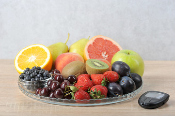 Acceptable fruits for diabetes. On a plate are apples, pears, kiwi, grapefruit, plums, blueberries, strawberries, cherries, orange, peaches. Next to a plate is a glucometer. Light background. Close-up