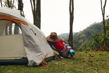 A backpack with a mat and a hat near to a tent on a grass in a camping on a background of mountains and forest.