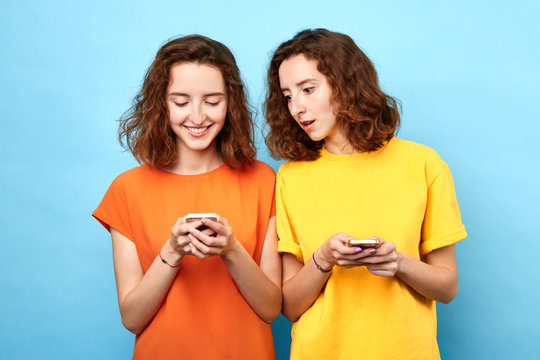 Two Identical Twins Holding Mobile Phones, Making A Phone Call, Playing Online Games, Surfing, Browsing The Net . Isolated Blue Background, Studio Shot