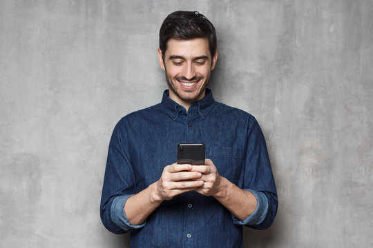 Smiiling business man in denim shirt and trendy eyeglasses standing against gray textured wall, typing a message using his phone