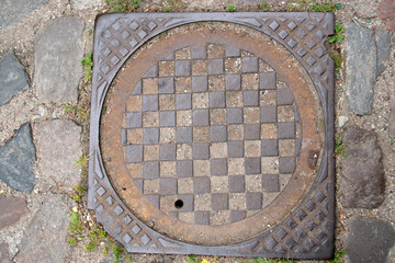 The well of the city drainage sewer with a metal corrugated brown lid with a chessboard pattern with a chipped corner, among the gray stones of the pavement