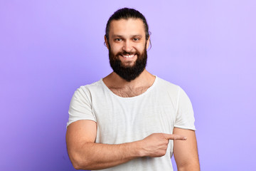 Portrait of a happy young man in white t-shirt pointing finger away at copy space isolated over...