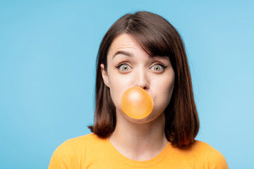 Young amazed brunette staring at you while blowing chewing-gum bubble