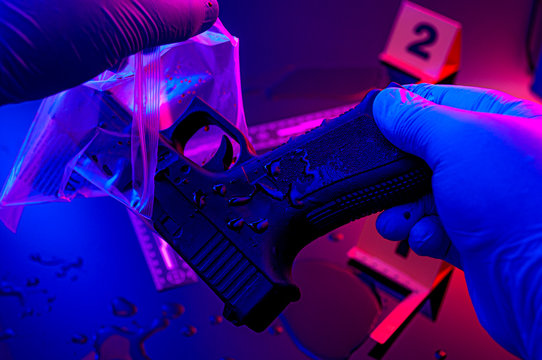 Forensic science, murder weapon and criminal investigation concept theme with detective wearing latex gloves bagging gun to send to lab in a dark crime scene illuminated by red and blue cop car lights