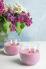 Obraz na płótnie Canvas Burning wax candles in glass holders and lilac flowers on white table, space for text