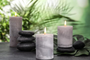 Fototapeta na wymiar Burning candles and spa stones on grey table against blurred green background