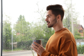 Fototapeta premium Happy handsome man with cup of coffee near window on rainy day