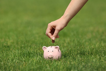 Young woman putting coin into piggy bank on green grass outdoors, closeup