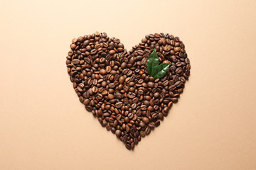 Heart shaped pile of coffee beans and fresh green leaves on light orange background, top view