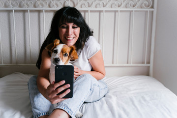 young caucasian woman on bed taking a selfie with mobile phone with her Cute small dog. Love for...
