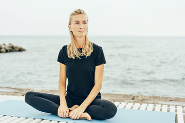 Fototapeta na wymiar A young slender woman sits in a lotus position after a basic sports training on the bay. Portrait of a beautiful blonde with freckles on her face, natural beauty.