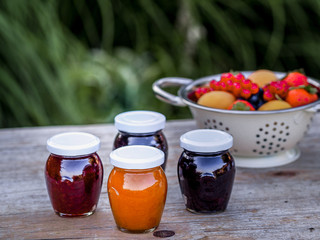 Four jars with homemade jam from blackberries, strawberries and apricots on the table in the garden. Bowl of fresh fruits on the background.