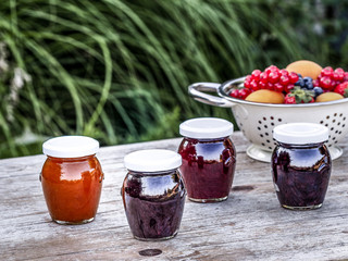 Four jars with homemade jam from blackberries, strawberries and apricots on the table in the garden. Bowl of fresh fruits on the background.