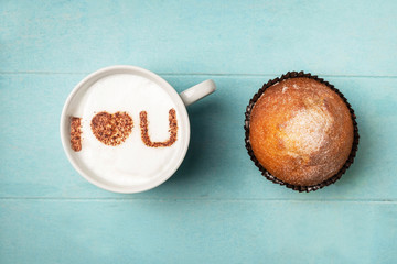 White cup of coffee with the inscription on the foam i love you and a cupcake on a blue wooden background, top view, copy space. Love confession concept, heart symbol.