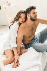 Naklejka premium sexy couple sitting in bed and holding hands while girl looking at camera and man looking away