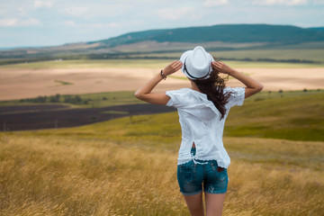 A young woman stands with her back to the frame, looks at the mountains, holds her hat with her hands.
