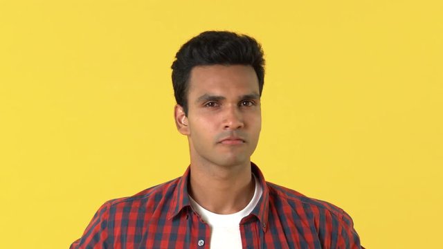 Handsome young Indian man in plaid shirt saying no by shaking head against yellow background