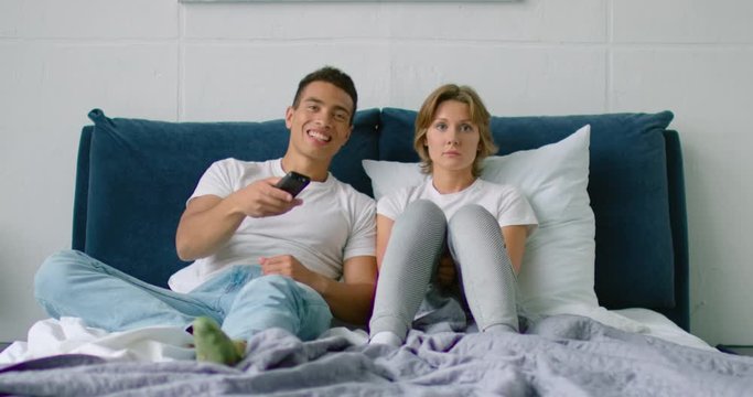 Young mixed couple is watching TV, lying in bed at home, man is interested, watching his favorite TV show, film, holding remote controller, woman is bored. Caucasian, white. 4K, shot on RED camera.