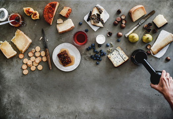 Wine snack variety. Flat-lay of cheeses, honey, fruit, nuts, crackers, female hands pouring wine from bottle to glass over grey concrete background, top view, copy space. Gathering, party food concept