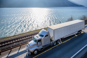 Big rig semi truck transporting cargo in dry van semi trailer running on sunny road along wide river