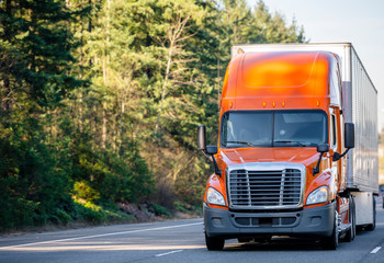 Orange big rig semi truck transporting cargo in semi trailer running on the road with trees - Powered by Adobe