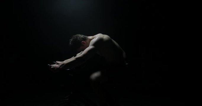Side Shot Of Man Exercising On Exercise Bike Shirtless Training As Smoke Falls On Him In A Dark Background